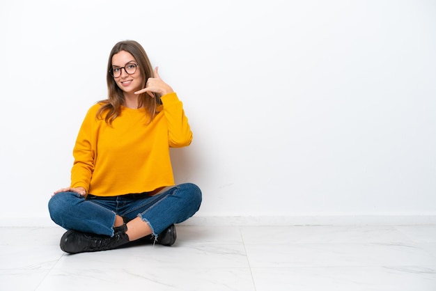 Jonge blanke vrouw zittend op de vloer geïsoleerd op een witte achtergrond telefoon gebaar maken Bel me terug teken