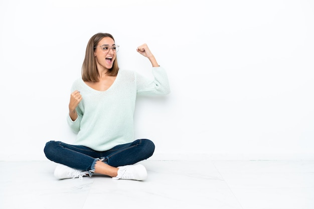 Jonge blanke vrouw zittend op de vloer geïsoleerd op een witte achtergrond vieren een overwinning