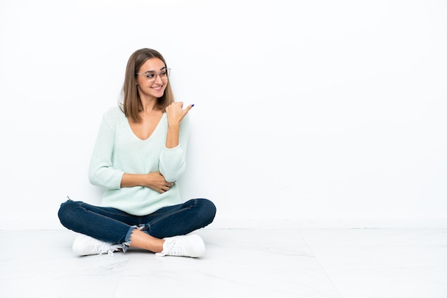 Jonge blanke vrouw zittend op de vloer geïsoleerd op een witte achtergrond wijzend naar de zijkant om een product te presenteren