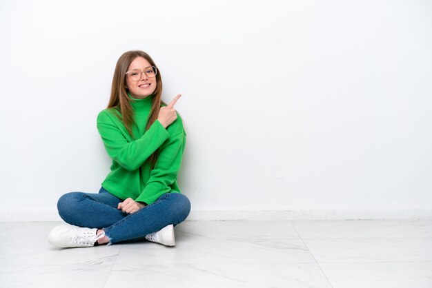 Jonge blanke vrouw zittend op de vloer geïsoleerd op een witte achtergrond wijzend naar de zijkant om een product te presenteren