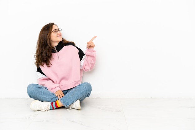 Jonge blanke vrouw zittend op de vloer geïsoleerd op een witte achtergrond wijzende vinger naar de zijkant en een product presenteren