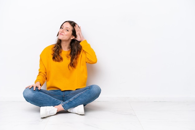 Jonge blanke vrouw zittend op de vloer geïsoleerd op een witte muur luisterend naar iets door hand op het oor te leggen