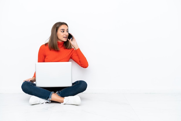Jonge blanke vrouw zittend op de vloer met een laptop die een gesprek voert met de mobiele telefoon