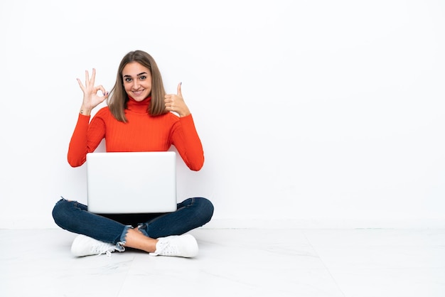 Jonge blanke vrouw zittend op de vloer met een laptop met ok teken en duim omhoog gebaar