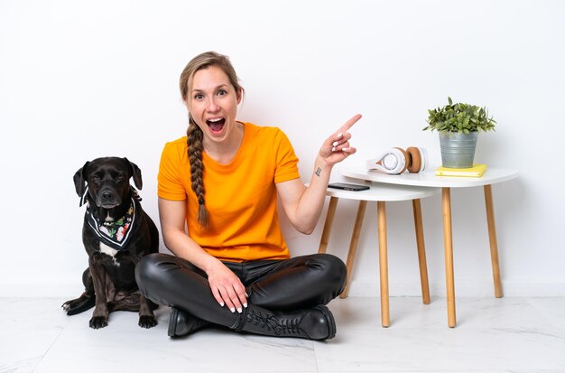 Jonge blanke vrouw zittend op de vloer met zijn puppy geïsoleerd op een witte achtergrond verrast en wijzende vinger naar de zijkant