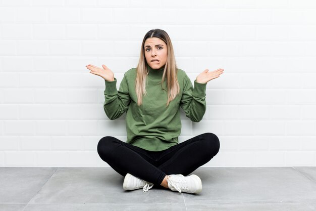 Jonge blanke vrouw zittend op de vloer verward en twijfelachtig schouders ophalen om een kopie ruimte te houden.