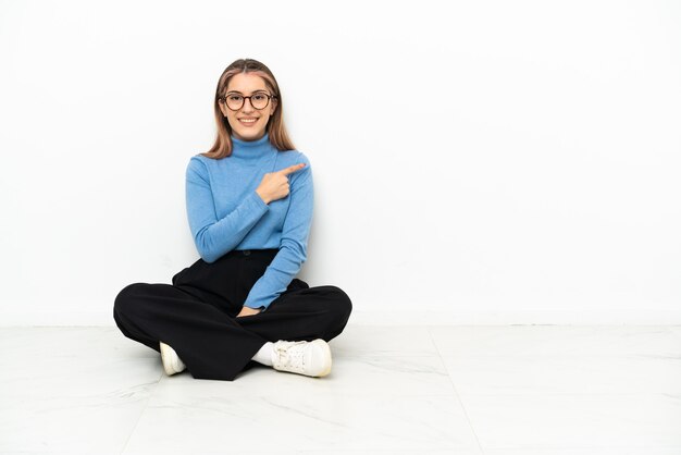 Jonge blanke vrouw zittend op de vloer wijst naar de zijkant om een product te presenteren