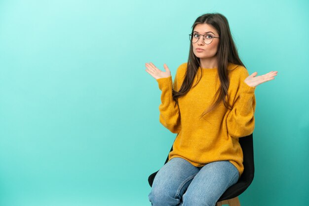 Jonge blanke vrouw zittend op een stoel geïsoleerd op een blauwe achtergrond die twijfelt terwijl ze haar handen opsteekt