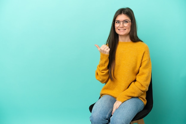 Jonge blanke vrouw zittend op een stoel geïsoleerd op een blauwe achtergrond, wijzend naar de zijkant om een product te presenteren