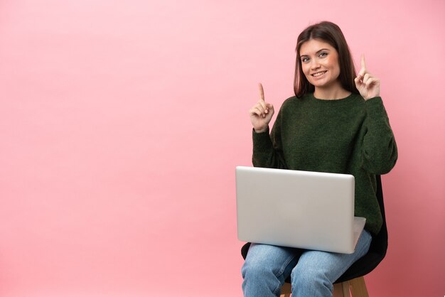 Jonge blanke vrouw zittend op een stoel met haar laptop geïsoleerd op een roze achtergrond die een geweldig idee benadrukt great