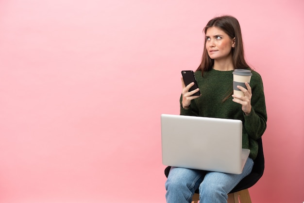 Jonge blanke vrouw zittend op een stoel met haar laptop geïsoleerd op roze achtergrond koffie te houden om mee te nemen en een mobiel terwijl ze iets denkt