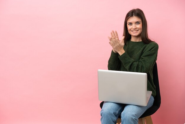 Jonge blanke vrouw zittend op een stoel met haar laptop geïsoleerd op roze muur applaudisseren na presentatie in een conferentie