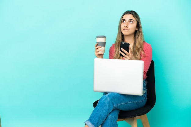 Jonge blanke vrouw zittend op een stoel met haar pc geïsoleerd op blauwe achtergrond met koffie om mee te nemen en een mobiel terwijl ze iets denkt thinking