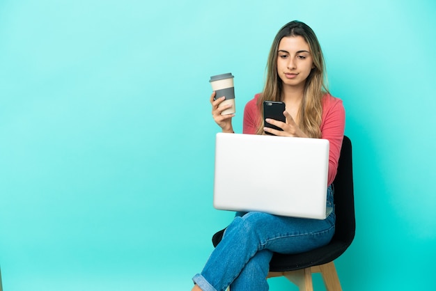 Jonge blanke vrouw zittend op een stoel met haar pc geïsoleerd op blauwe achtergrond met koffie om mee te nemen en een mobiel