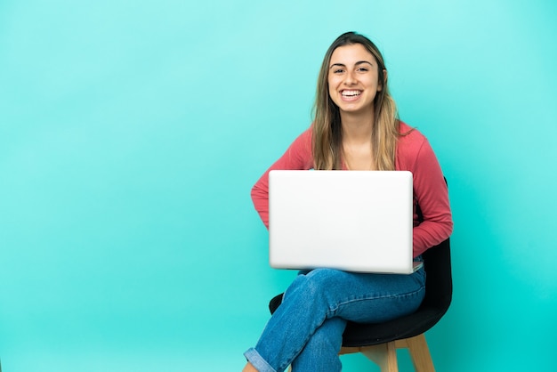 Jonge blanke vrouw zittend op een stoel met haar pc geïsoleerd op blauwe achtergrond poseren met armen op heup en glimlachen