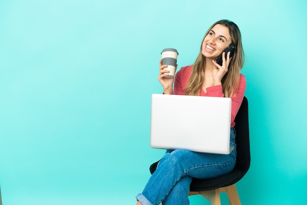 Jonge blanke vrouw zittend op een stoel met haar pc geïsoleerd op blauwe muur met koffie om mee te nemen en een mobiel