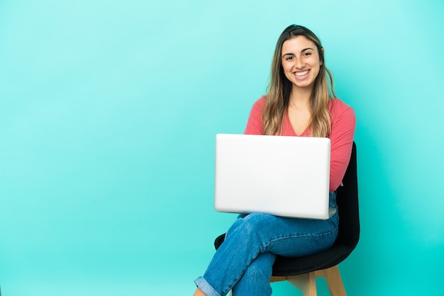 Jonge blanke vrouw zittend op een stoel met haar pc geïsoleerd op een blauwe achtergrond en houdt de armen gekruist in frontale positie