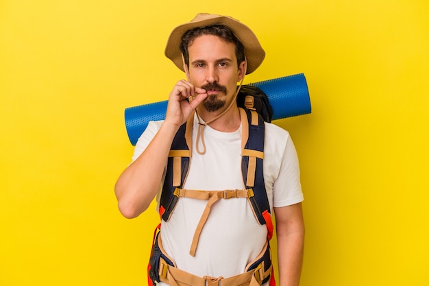 Jonge blanke wandelaar man geïsoleerd op gele achtergrond met vingers op lippen die een geheim houden.