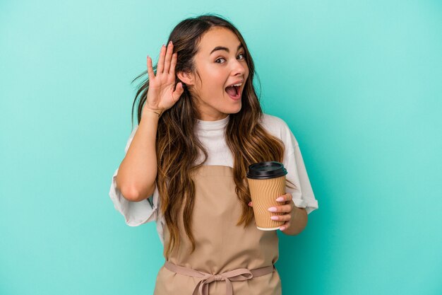 Jonge blanke winkelbediende vrouw met een afhaalmaaltijden koffie geïsoleerd op blauwe achtergrond probeert te luisteren naar een roddel.