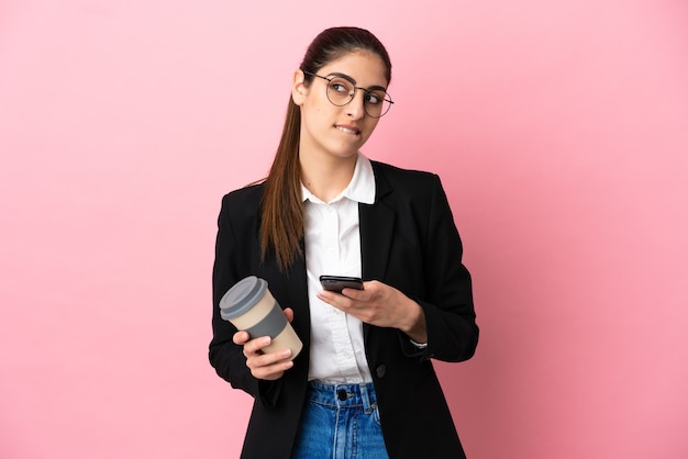 Jonge blanke zakenvrouw geïsoleerd op roze achtergrond met koffie om mee te nemen en een mobiel terwijl ze iets denkt while