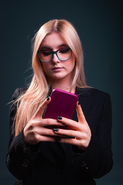 Jonge blanke zakenvrouw met behulp van een mobiele telefoon met een zwarte blazer en bril in een fotostudio met een blauwe donkere achtergrond.