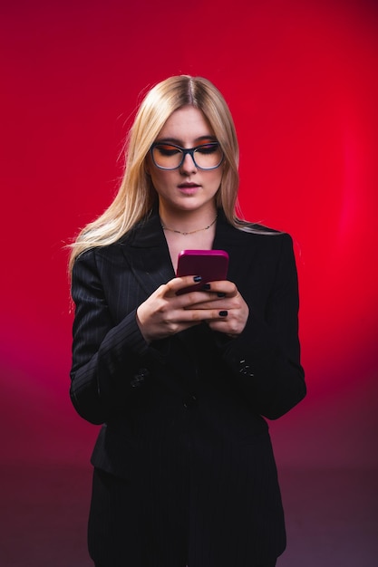 Jonge blanke zakenvrouw met behulp van een mobiele telefoon met een zwarte blazer en bril in een fotostudio met een rode donkere achtergrond.