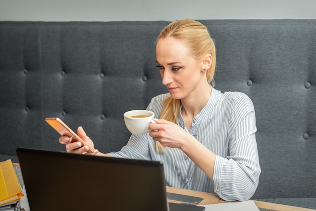 Jonge blanke zakenvrouw met smartphone en kopje koffie thuis kantoor werken
