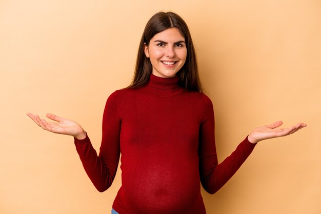 Jonge blanke zwangere vrouw geïsoleerd op beige achtergrond met een welkome uitdrukking.