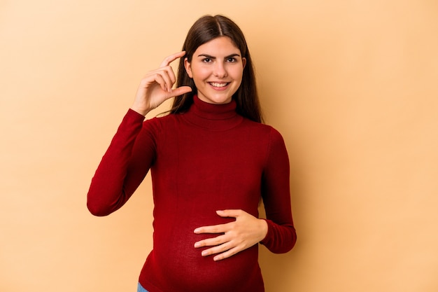 Jonge blanke zwangere vrouw geïsoleerd op beige achtergrond met iets kleins met wijsvingers, glimlachend en zelfverzekerd.