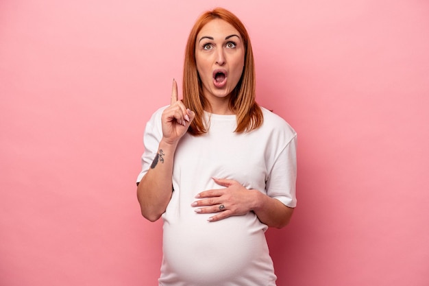 Jonge blanke zwangere vrouw geïsoleerd op roze achtergrond wijzend ondersteboven met geopende mond.