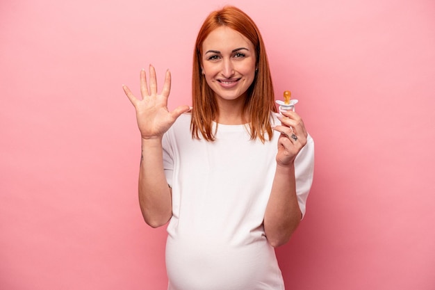 Jonge blanke zwangere vrouw met fopspeen geïsoleerd op een roze achtergrond glimlachend vrolijk met nummer vijf met vingers