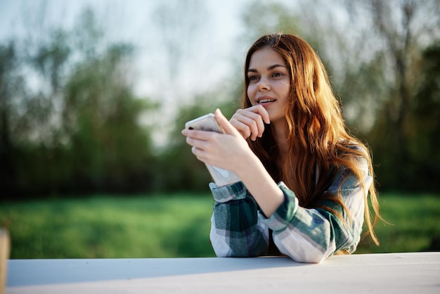 Jonge blogger-freelancer die haar telefoon vasthoudt terwijl ze ernaar kijkt en buitenshuis in een groen park werkt en peinzend online naar het scherm kijkt