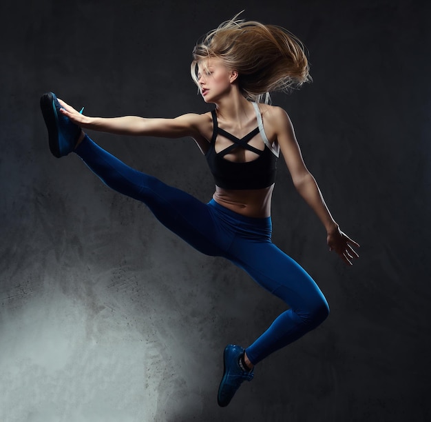 Jonge blonde ballerina in sportkleding danst en springt in een studio. Geïsoleerd op een donkere achtergrond.