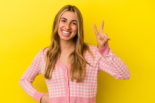 Jonge blonde blanke vrouw die op gele muur wordt geïsoleerd die nummer twee met vingers toont