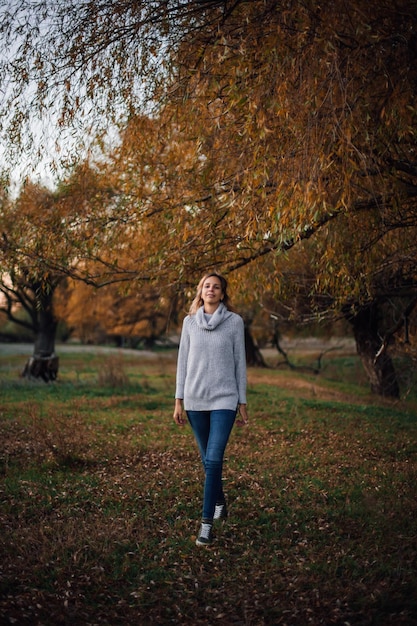 Jonge blonde blanke vrouw die rechtdoor loopt en naar de camera kijkt glimlachend in een grijze trui, spijkerbroek en sneakers met bomen bedekt met zonsondergang op de achtergrond Herfstwandeling in het bos