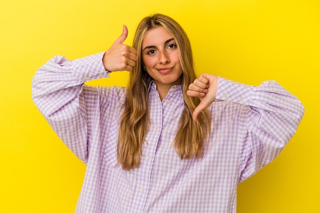 Jonge blonde blanke vrouw geïsoleerd op gele achtergrond duimen opdagen en duimen naar beneden, moeilijk kiezen concept