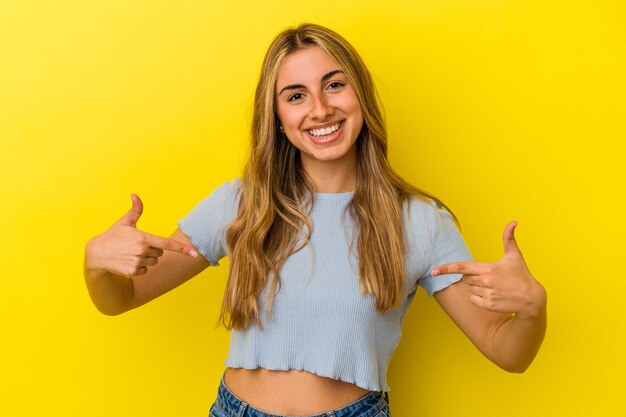 Jonge blonde blanke vrouw geïsoleerd op gele muur persoon met de hand wijzend naar een shirt kopie ruimte, trots en zelfverzekerd