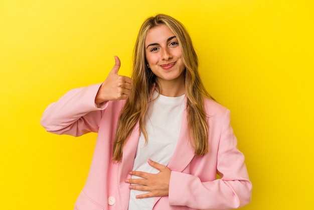 Jonge blonde blanke vrouw geïsoleerd op gele muur raakt buik, glimlacht zachtjes, eten en tevredenheid concept.