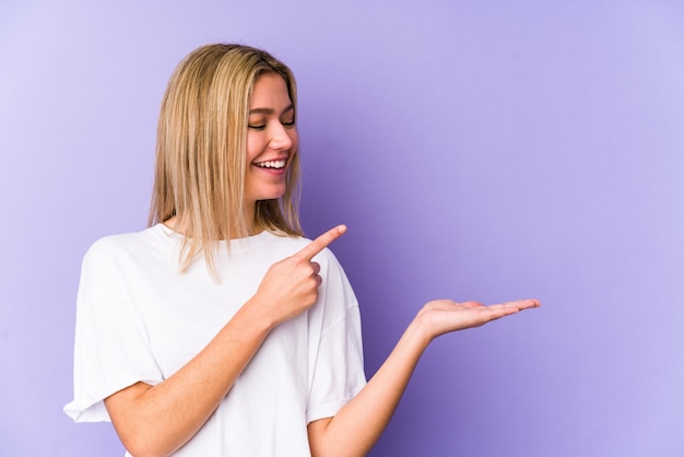 Jonge blonde blanke vrouw geïsoleerd opgewonden met een kopie ruimte op de palm.