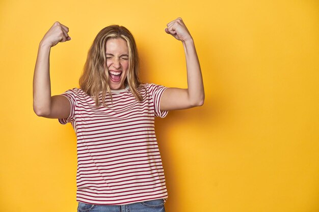 Foto jonge blonde blanke vrouw in een rood gestreepte t-shirt op een gele achtergrond die de vuist heft na een overwinningsconcept