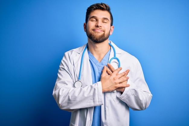 Jonge blonde dokter man met baard en blauwe ogen in witte jas en stethoscoop glimlachend met de handen op de borst met gesloten ogen en dankbaar gebaar op het gezicht Gezondheidsconcept