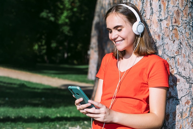 Jonge blonde draagt een rood shirt kijkt naar de mobiele telefoon en luistert naar muziek in een park