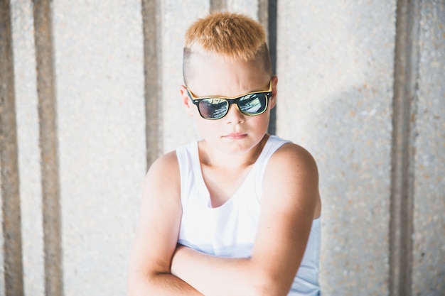 Jonge blonde jongen met zonnebril staat tegen de muur