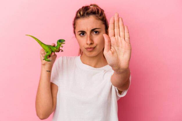 Jonge blonde kaukasische vrouw die een dinosaurusminiatuur houdt die op roze muur wordt geïsoleerd die zich met uitgestrekte hand bevindt die stopbord toont, dat u verhindert