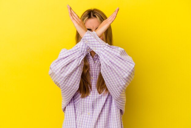 Jonge blonde Kaukasische vrouw die op gele muur wordt geïsoleerd die twee gekruiste wapens houdt, ontkenningsconcept.