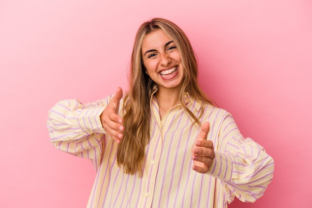 Jonge blonde kaukasische vrouw die op roze muur wordt geïsoleerd die iets met beide handen, productpresentatie houdt