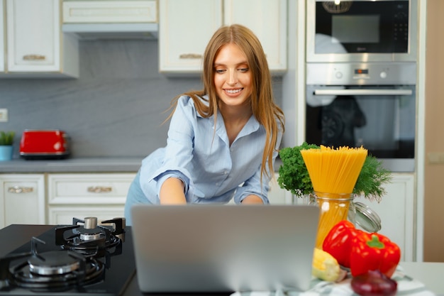 Jonge blonde Kaukasische vrouw met lang haar die laptop in een keuken met behulp van