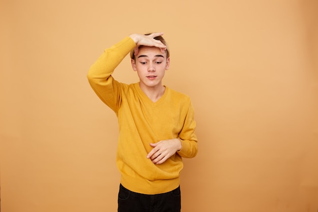 Jonge blonde man gekleed in gele trui houdt zijn vingers boven zijn wenkbrauwen op de beige achtergrond in de studio.