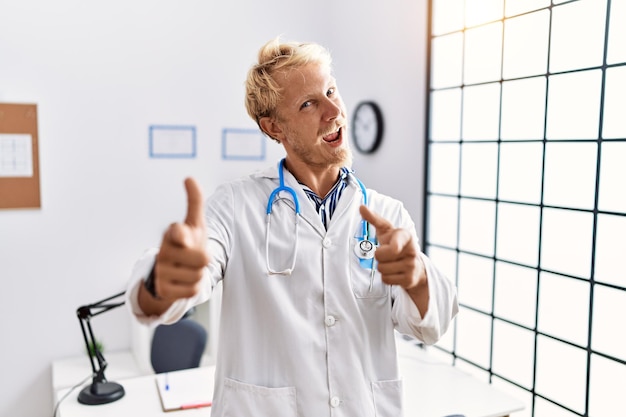 Jonge blonde man in doktersuniform en stethoscoop in kliniek wijzende vingers naar camera met blij en grappig gezicht, goede energie en vibes