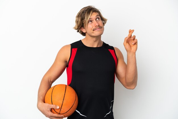 Jonge blonde man met een bal van basketbal geïsoleerd op een witte muur met vingers kruisen en het beste wensen